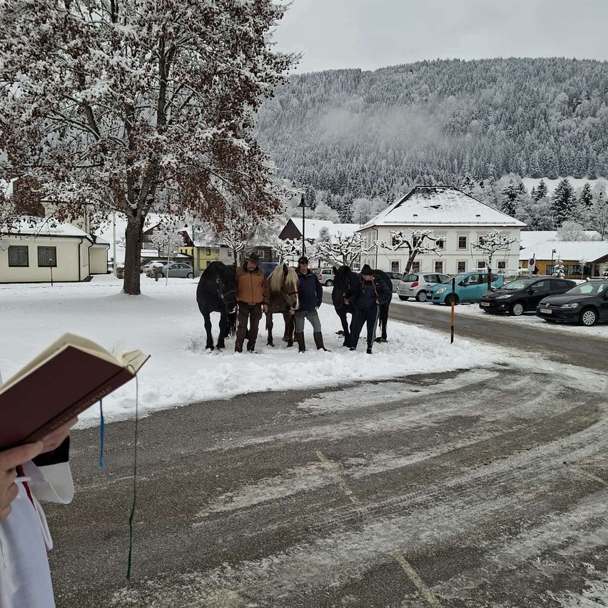 Ein Priester hält ein Buch vor einer Kirche an einem verschneiten Tag. Mehrere Menschen und Pferde stehen im Schnee. Im Hintergrund befinden sich Gebäude und schneebedeckte Berge.