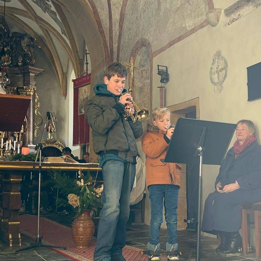 Zwei junge Jungen spielen Trompeten in einer Kirche. Eine ältere Frau beobachtet sie von einem Holzstuhl aus. Ein Tisch mit einer Kerze und Blumen steht hinter ihnen.