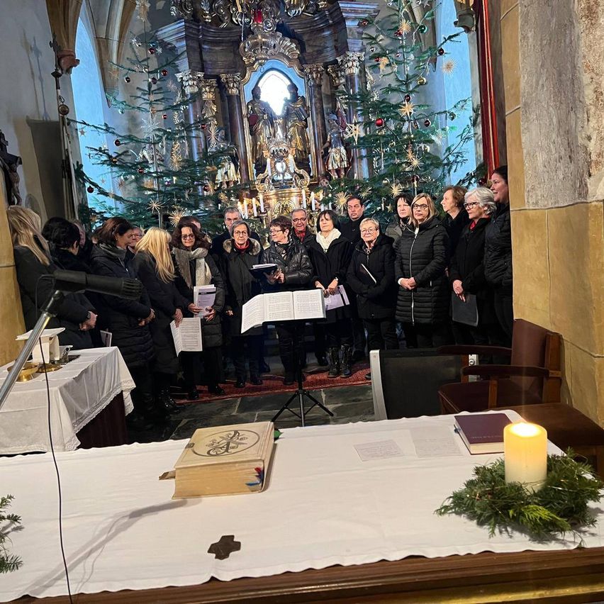 Ein Chor steht in einer Kirche mit Weihnachtsdekoration. Eine Kerze ist angezündet, und ein Buch liegt auf dem Altar. Einige Leute halten ein Notenblatt.