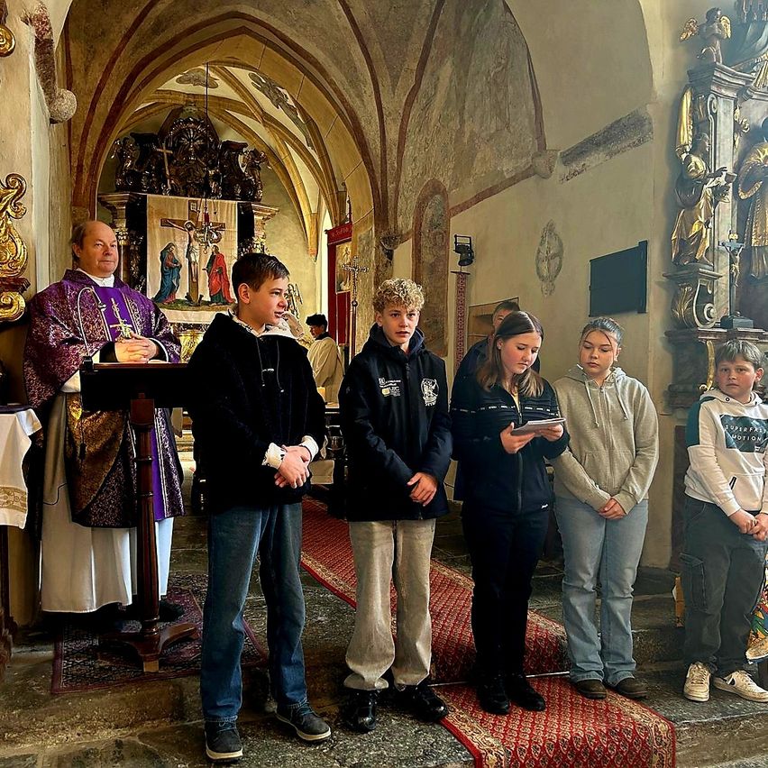 Ein Priester steht vor einem Pult in einer Kirche mit vier Kindern, die vor ihm stehen und aus einem Buch lesen.