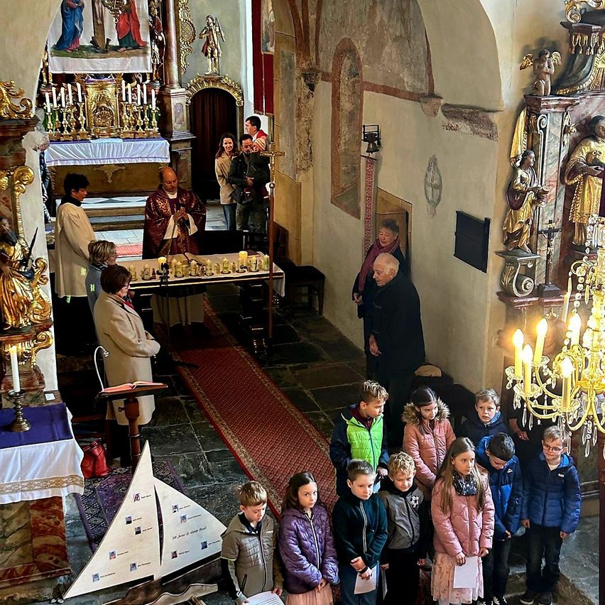 Eine Gruppe von Kindern steht in einer Kirche und hört einem Priester zu. Altar mit Kerzen und ein Segelboot auf dem Boden. An den Wänden befinden sich religiöse Gemälde und Statuen.