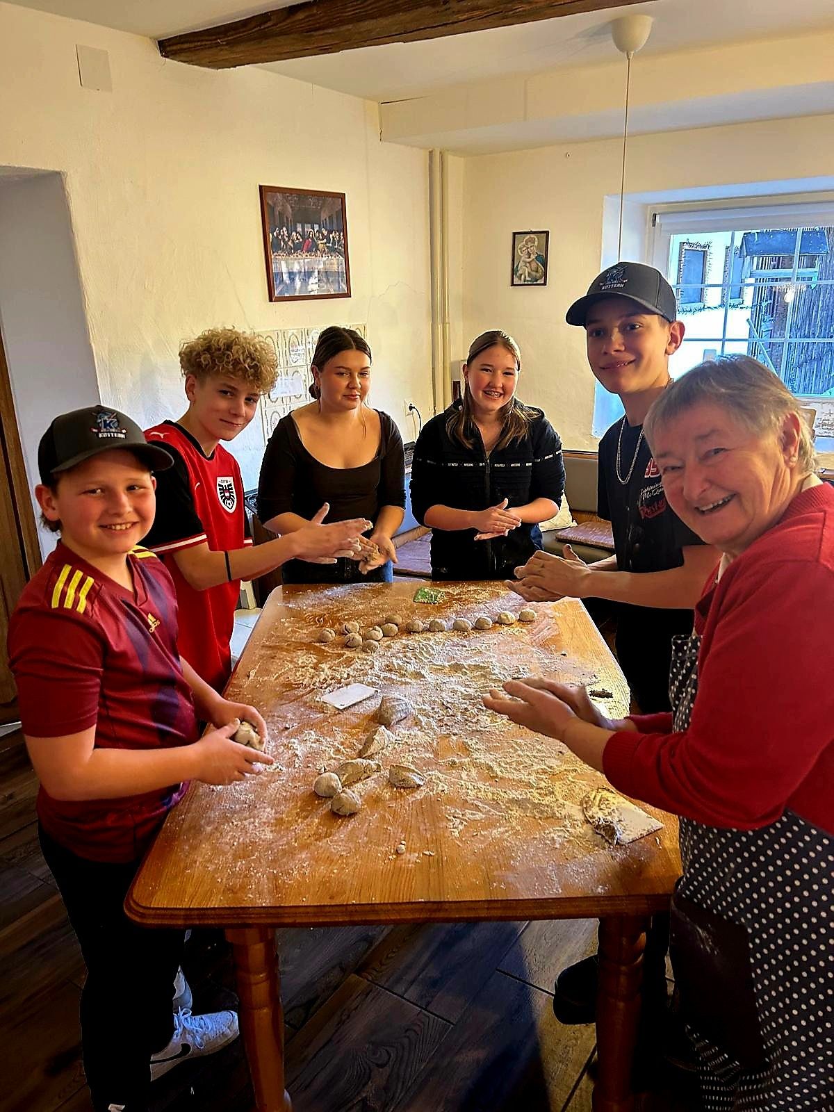 Eine Gruppe von Menschen ist in einer Küche und macht Teig auf einem Tisch. Zwei Frauen lächeln, während zwei Männer in die Kamera schauen.
