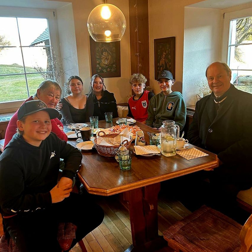 Eine Familie sitzt lächelnd um einen Esstisch herum. Auf dem Tisch stehen Tassen, Teller und Körbe. Der Raum hat große Fenster und einen Bilderrahmen an der Wand.