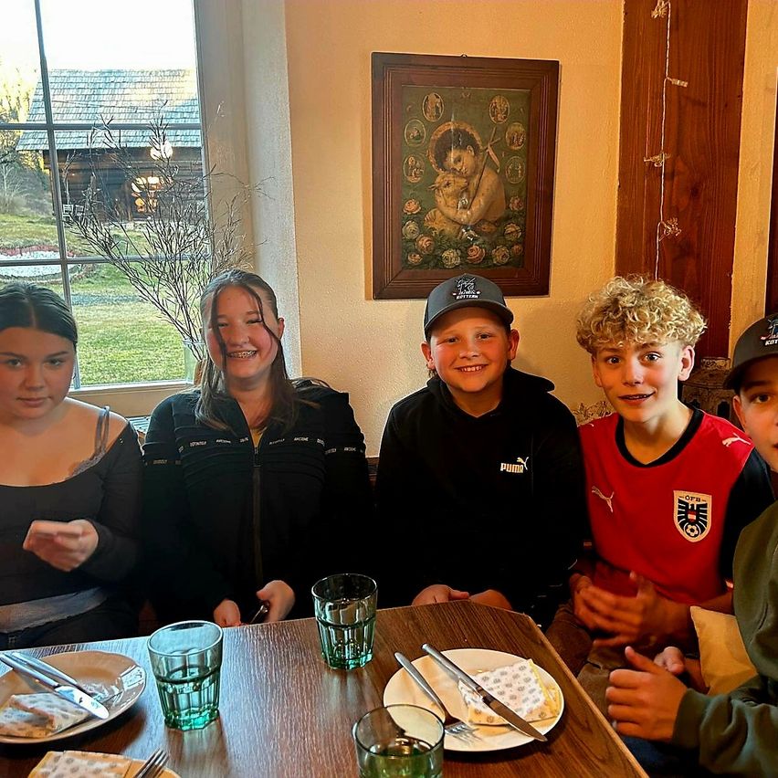 Fünf junge Menschen sitzen lächelnd um einen Tisch im Inneren. Auf dem Tisch stehen Teller, Gläser und Besteck. Hinter ihnen zeigt ein Fenster eine Rasenlandschaft und ein Haus. Ein Bilderrahmen und dekorative Lichter schmücken die Wand.