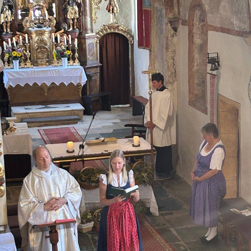 Ein Priester und eine Frau in traditioneller Kleidung beten in einer Kirche, während ein anderer Priester bei einem Kreuz steht. Der Altar ist mit Kerzen und Blumen geschmückt.