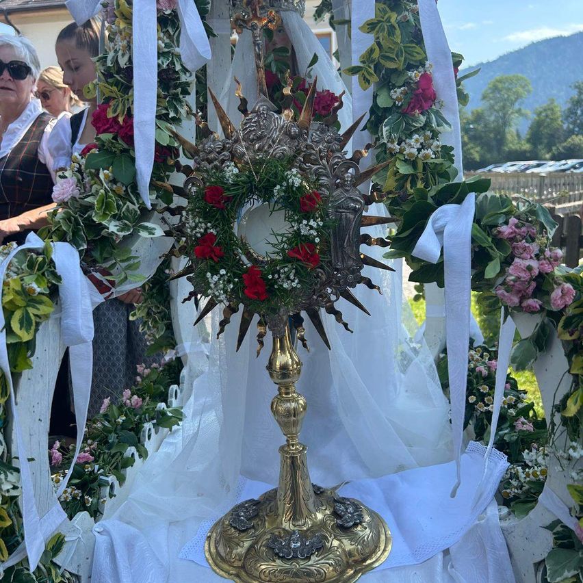 Ein goldener und silberner Monstranz, geschmückt mit roten Rosen und grünen Blättern, umgeben von weißen Bändern und Blumenarrangements, mit Menschen in traditioneller Kleidung im Hintergrund.