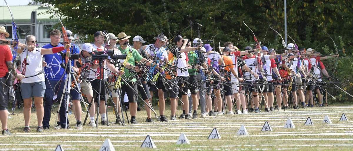 Eine Gruppe von Bogenschützen steht in einer Reihe, mit Bögen und Pfeilen, auf einem Rasenfeld mit nummerierten Markierungen. Bäume und eine Straßenlaterne im Hintergrund.