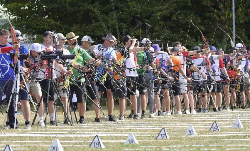 A group of archers stand in a line, holding bows and arrows, on a grassy field with numbered markers. Trees and a streetlight are in the background.