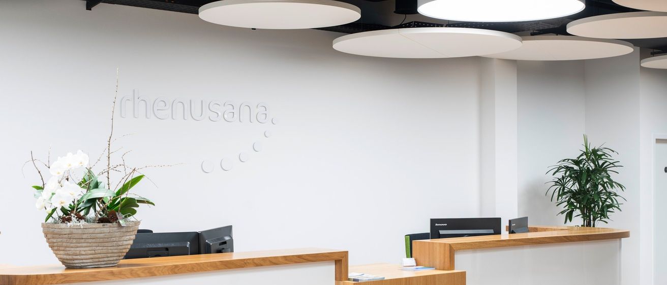 Ein modernes Büro mit weißen Wänden, Holztheken und runden Deckenleuchten. Auf der linken Theke steht eine Pflanze, auf der rechten ein Computermonitor. Das Wort 'rhenusana' ist an der Wand geschrieben.