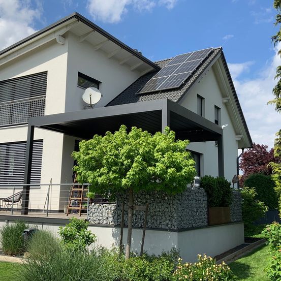 Ein modernes Haus mit Solarpanelen auf dem Dach und einer schattigen Terrasse davor. Das Haus hat eine Steinmauer und einen Baum davor.