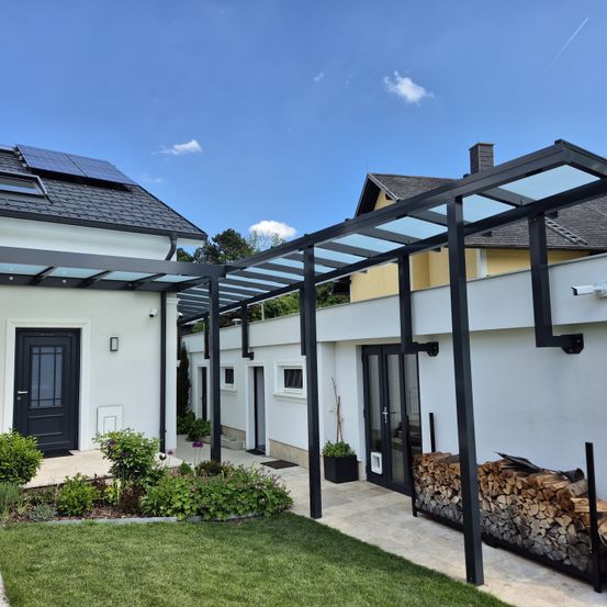 Ein modernes Haus mit Solarmodulen auf dem Dach, einer Glaspergola und einem gepflegten Garten mit einem Holzstapel.