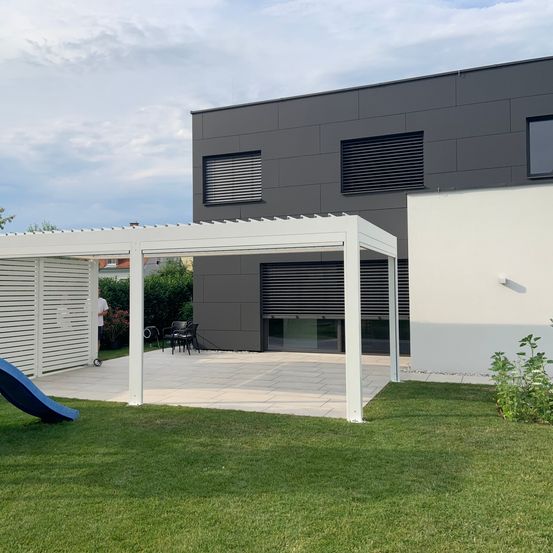 Ein modernes Haus mit einer weißen Pergola im Garten, mit einer Terrasse, Stühlen und einem Rutschvergnügen auf dem Rasen.