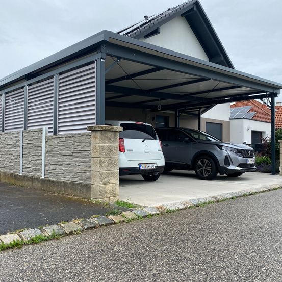 Zwei Autos in einer modernen Garage mit Metalldach und Steinwänden unter einem bewölkten Himmel geparkt.