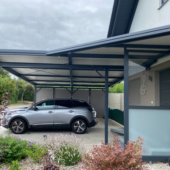 Ein silberner SUV ist unter einem Metallcarport vor einem Haus geparkt. Der Carport hat eine Glaswand und einen Metallrahmen.