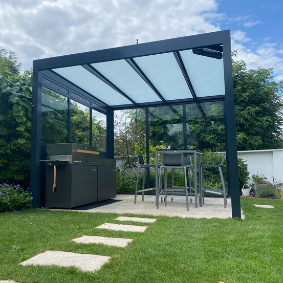 Eine moderne Outdoor-Terrasse mit einem Glaspavillon, der einen Grill, einen Tisch mit Stühlen und einen Gehweg mit Trittsteinen enthält. Der Bereich ist von Grünpflanzen umgeben.