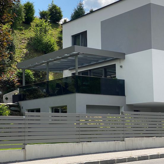 Ein modernes weißes Haus mit einem grauen Balkon hat ein Glasgeländer und eine überdachte Terrasse mit einem grauen Dach.