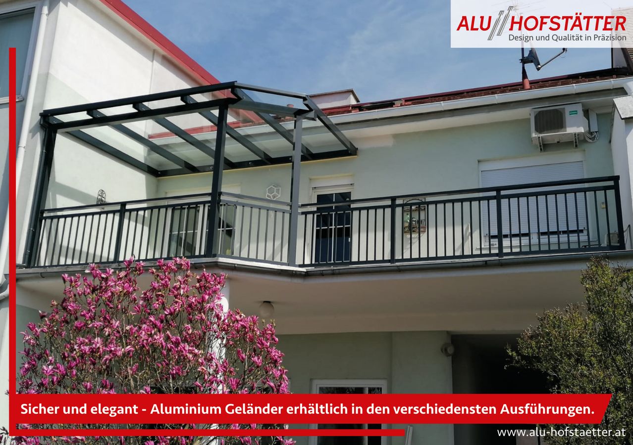 Ein weißes Gebäude mit einem Balkon, der mit einem schwarzen Aluminiumgeländer ausgestattet ist. Der Balkon hat ein Glasdach und ist mit rosa Blumen davor geschmückt. Das Bild enthält einen Wasserzeichen mit dem Text 'ALU//HO Design u'.
