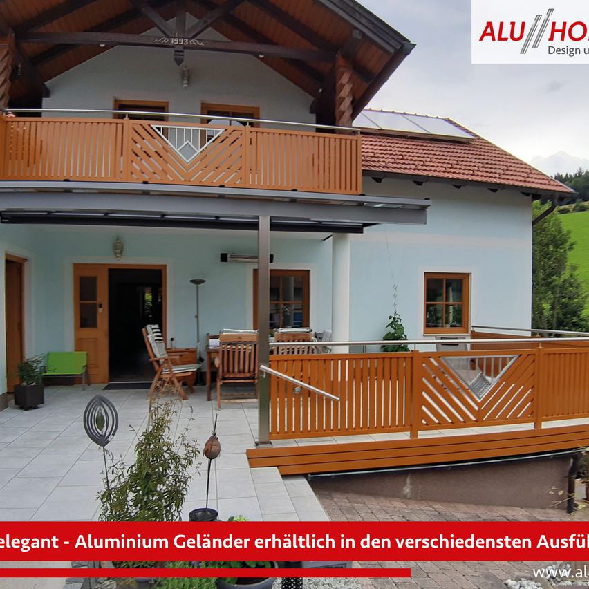 Das Bild zeigt ein modernes Haus mit Aluminiumgeländern auf dem Balkon und der Terrasse. Das Haus hat ein Ziegeldach und Holzakzente. Der Balkon hat ein geometrisches Design und die Terrasse ist mit Holzstühlen, einem Tisch und Topfpflanzen ausgestattet. Das Haus ist von Grün umgeben.