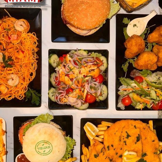 Eine Auswahl an Gerichten von Miami Pizzeria & Ristorante, einschließlich Pasta, Burger, Salate und frittierte Speisen, ist auf schwarzen Tellern zu sehen.