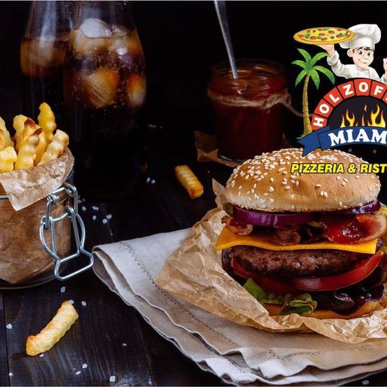 Ein Burger mit mehreren Schichten von Toppings, einschließlich Käse, Tomate und Speck, sitzt auf einem Papierumschlag auf einem Holztisch. Im Hintergrund befinden sich Gläser mit Limonade, ein Glas mit Sauce und ein Korb mit Pommes frites. Das Bild zeigt ein Logo für Holzofen Miami, ein Pizzeria und Restaurant.