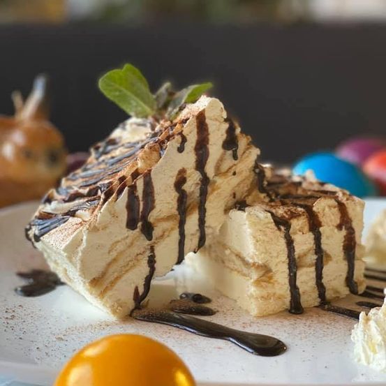 Ein Stück Tiramisu-Dessert mit Schokoladensauce und Minzblatt auf einem weißen Teller, mit Ostereiern im Hintergrund.