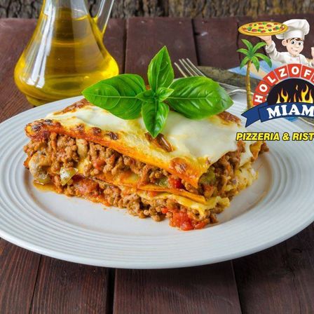 Ein Stück Lasagne mit geschmolzenem Käse und Basilikumblättern auf einem weißen Teller, mit einer Ölflasche und einer Gabel in der Nähe. Das Bild zeigt ein Logo für Holzofen Miami.