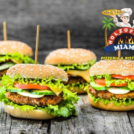 Vier Burger mit Salat, Tomaten und Gewürzgurken auf einem Holztisch mit einem Logo für Holzofen Miami Pizzeria & Ristorante im Hintergrund.