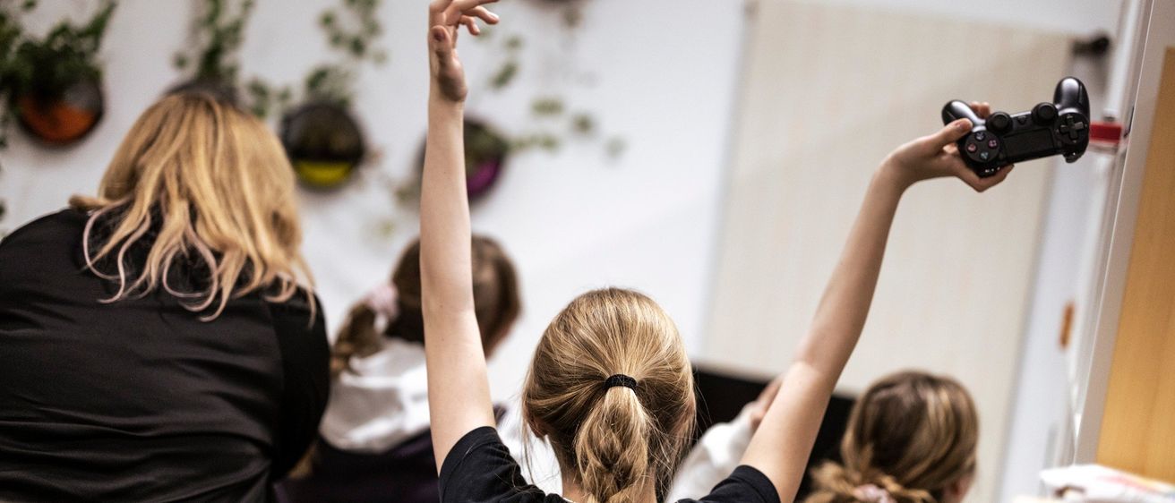 Ein Mädchen mit blonden Haaren, das ein schwarzes T-Shirt trägt, hebt die Hände hoch und hält einen Controller.