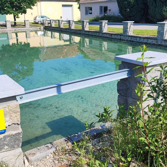 Ein klarer Kanal mit Steinsäulen und einem Metallgeländer, umgeben von Grün und reflektiert die Gebäude und Bäume auf der anderen Seite.