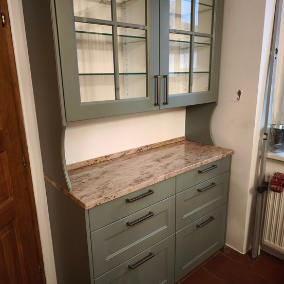 Eine Küche mit einem grünen Schrank mit Glastüren und Schubladen, einer Marmorarbeitsplatte und einer hölzernen Tür auf der linken Seite.
