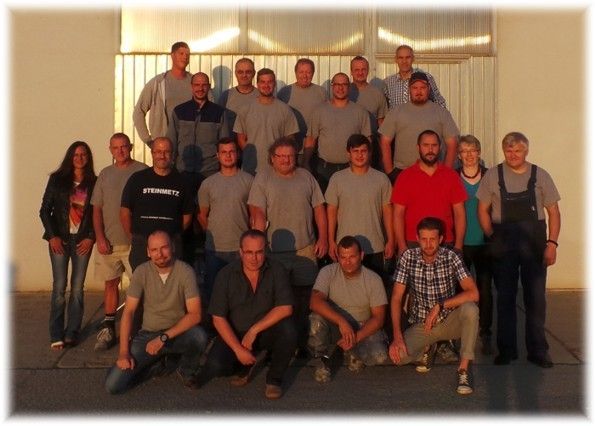 Eine Gruppe von Menschen, hauptsächlich Männer, posiert für ein Foto vor einem Gebäude. Sie tragen Freizeitkleidung, und einige tragen Brillen. Der Mann im roten Shirt sitzt auf dem Boden.