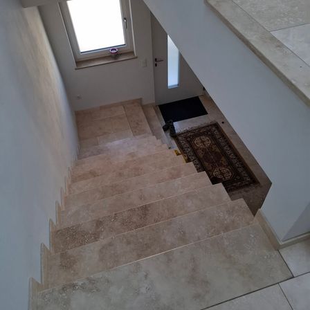 Eine moderne Treppe mit beigefarbenen Fliesen, die zu einer weißen Tür mit einem Fenster darüber führt. Ein Teppich liegt am Fuß der Treppe.