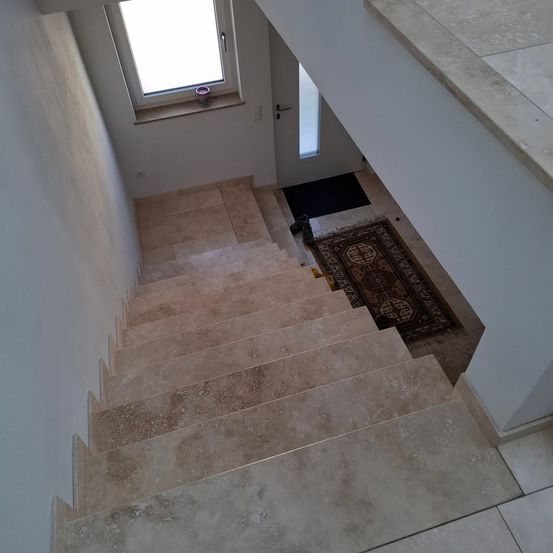Eine moderne Treppe mit beigefarbenen Fliesen, die zu einer weißen Tür mit einem Fenster darüber führt. Ein Teppich liegt am Fuß der Treppe.