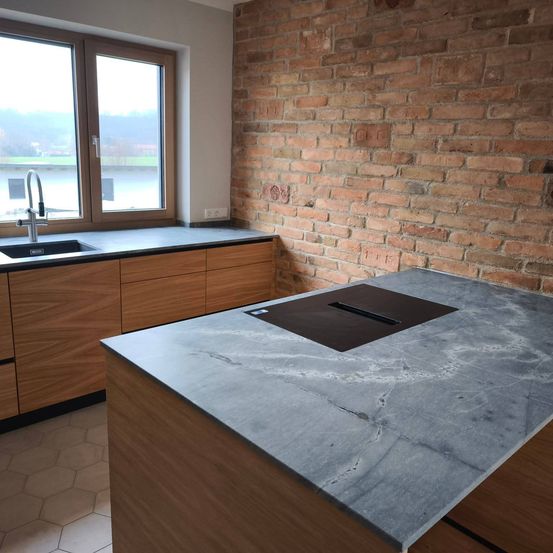 Eine moderne Küche mit einer großen Granitarbeitsplatte, Holzschränken und einer Ziegelwand. Ein Waschbecken und eine Armatur sind in der Nähe des Fensters zu sehen.