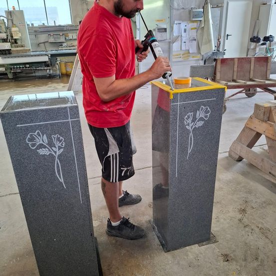 Ein Mann in einem roten T-Shirt und schwarzen Shorts trägt Versiegelung auf ein graues Monument mit Blumendesign auf. Der Schauplatz scheint ein Workshop zu sein.