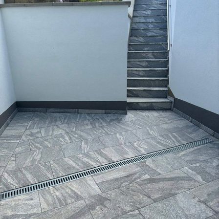 Ein grauer Steinterrasse mit einem Abfluss, der hindurchläuft, neben einer weißen Wand und einer Treppe.