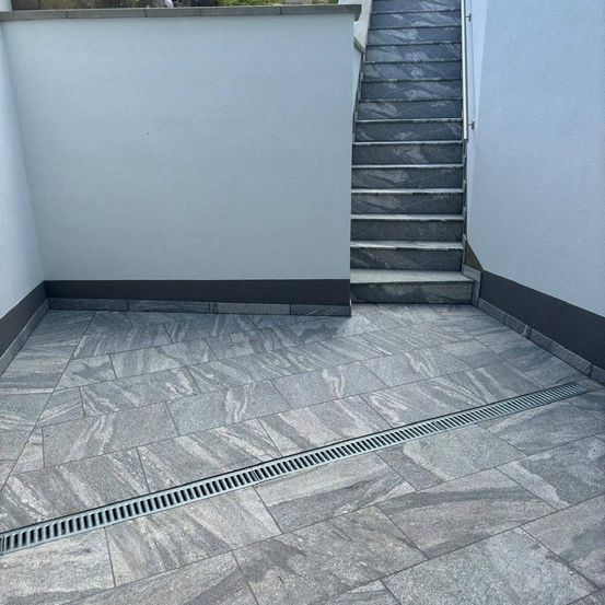 Ein grauer Steinterrasse mit einem Abfluss, der hindurchläuft, neben einer weißen Wand und einer Treppe.