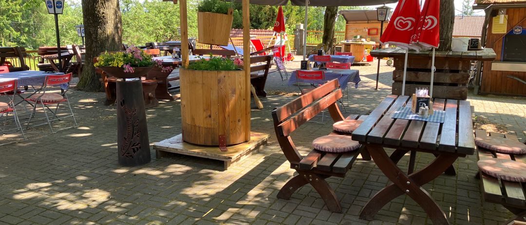 Außenbereich mit einem hölzernen Wasserbrunnen und Tischen unter Sonnenschirmen, in einer parkähnlichen Umgebung mit Bäumen, Blumen und Holzbänken.