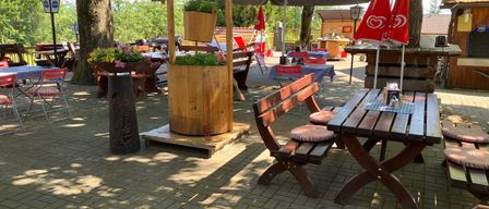 Außenbereich mit einem hölzernen Wasserbrunnen und Tischen unter Sonnenschirmen, in einer parkähnlichen Umgebung mit Bäumen, Blumen und Holzbänken.