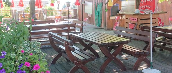 A sunlit outdoor restaurant with wooden tables, benches, and red umbrellas. Flowers are planted near the tables. Trees and plants provide shade.