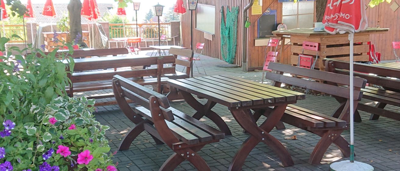 Ein sonniger Außenbereich eines Restaurants mit Holztischen, Bänken und roten Sonnenschirmen. Blumen stehen neben den Tischen. Bäume und Pflanzen bieten Schatten.