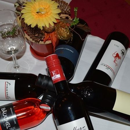 Fünf Weinflaschen und ein Weinglas stehen auf einem Tisch, mit einer roten Tischdecke und einem dekorativen Blumen-Centerpiece.