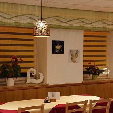 Ein Restaurant mit einer hölzernen Theke, zwei Tischen, Stühlen und Topfpflanzen. Es hängen Lampen an der Decke und Bilderrahmen an der Wand.