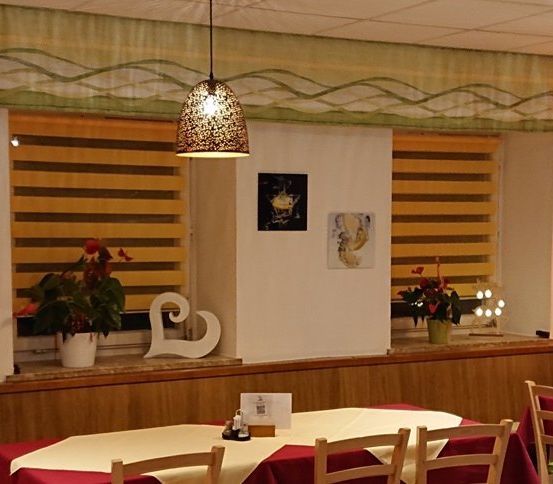 Ein Restaurant mit einer hölzernen Theke, zwei Tischen, Stühlen und Topfpflanzen. Es hängen Lampen an der Decke und Bilderrahmen an der Wand.