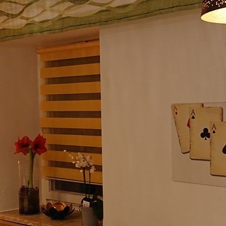 Ein Raum mit gelben Jalousien, einem Fenster und einer weißen Wand mit Spielkarten und einem Kreuz. An der Wand hängt eine Lampe.