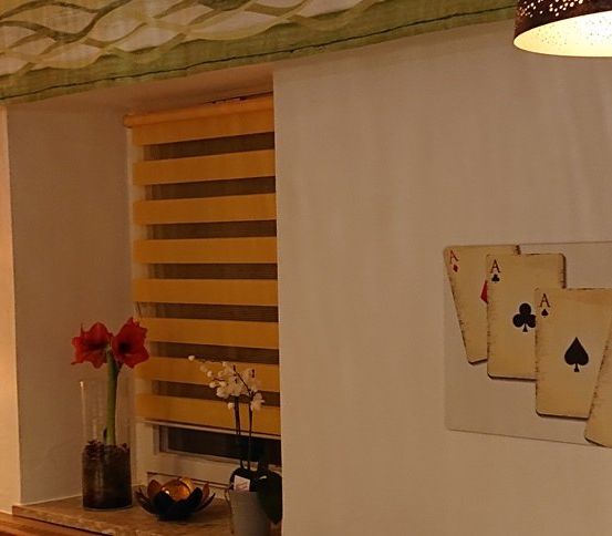 Ein Raum mit gelben Jalousien, einem Fenster und einer weißen Wand mit Spielkarten und einem Kreuz. An der Wand hängt eine Lampe.