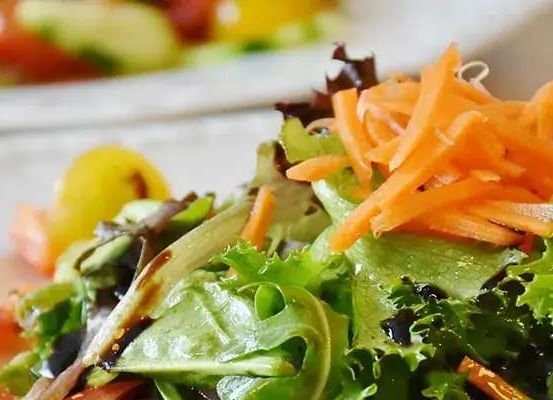 Ein Nahaufnahme eines frischen Salats auf einem weißen Teller mit geriebenen Karotten, Tomaten und grünem Blatt. Der Salat sieht leuchtend und appetitanregend aus und deutet auf eine gesunde Mahlzeit hin.