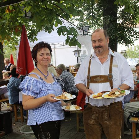 Zwei Personen in traditioneller bayerischer Kleidung halten Teller mit Essen. Ein Mann trägt braune Hosen und ein weißes Hemd mit Hosenträgern. Eine Frau trägt ein blaues Kleid mit weißen Punkten. Sie stehen in einem Außenbereich, wo andere Leute an Tischen sitzen.