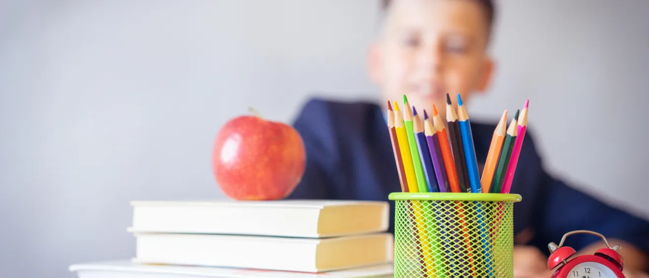 Ein junger Junge sitzt an einem Schreibtisch mit einem farbenfrohen Stifthalter, einer Brille und einem Stapel Bücher. Ein Apfel ruht auf den Büchern. Der Junge blickt mit einem leichten Lächeln nach vorne.