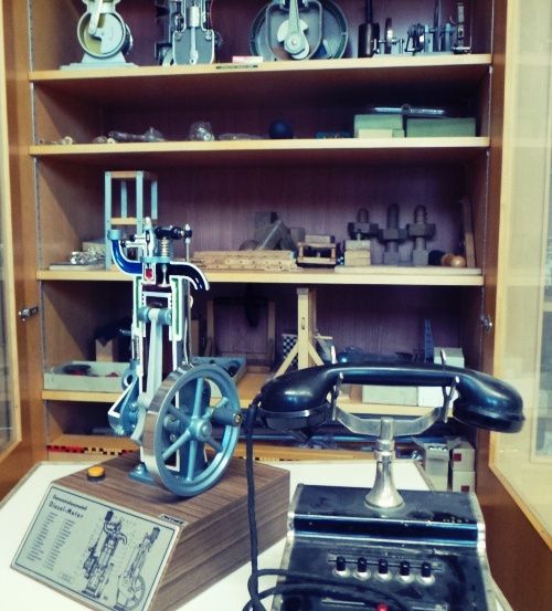Ein hölzerner Schrank zeigt verschiedene mechanische Modelle. Im Vordergrund sind ein Modell-Dampfmotor und ein alter Telefonapparat prominent ausgestellt. Der Schrank enthält zusätzliche mechanische Modelle, Werkzeuge und Teile.